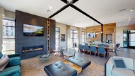 This elegantly designed two-floor amenity space blends contemporary architecture with warm, natural materials to create a refined and functional environment. The main level welcomes visitors with a striking marble-topped island counter positioned near steel-framed floor-to-ceiling windows that flood the space with natural light and offer sweeping views of the surrounding tree canopy and rolling hills beyond.

The open-plan layout seamlessly connects a lounge area, dining zone, and entertainment nook. A dramatic textured dark-tile accent wall anchors one end of the lounge, featuring a sleek linear gas fireplace beneath a wall-mounted display, complemented by a built-in bookcase and curated artwork. Warm wood-plank wall paneling adds contrast throughout, framing the dining area with its long communal table and blue upholstered seating beneath a globe pendant chandelier.

A dedicated coffee and refreshment station is outfitted with warm wood cabinetry, a beverage refrigerator, and a full coffee setup. A cozy media nook with built-in bench seating and a wood-clad accent wall provides an intimate viewing area.

The upper level offers a sun-drenched lounge with wraparound floor-to-ceiling glazing, delivering unobstructed panoramic views of the hillside landscape — an ideal setting for quiet retreat or casual gatherings. A staircase with sleek black metal railings connects both floors with understated elegance.
