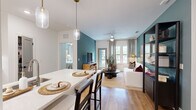 This thoughtfully designed 2-bedroom, 3-bathroom residence offers a functional and stylish single-floor layout. The open-concept living and kitchen area forms the heart of the home, where warm hardwood flooring and bold teal accent walls create a modern yet inviting atmosphere. The kitchen features white cabinetry, stainless steel appliances, a light quartz peninsula with bar seating, and pendant lighting — combining practicality with aesthetic appeal. The living area flows naturally from the kitchen, anchored by a ceiling fan, large windows, and a glass-paned exterior door that fill the space with natural light and views of the outdoors. A tall open shelving unit adds both storage and visual interest to the living room. The primary bedroom is a well-proportioned retreat with hardwood floors, a ceiling fan, large window with patterned drapes, and ample room for furnishings. The second bedroom is equally comfortable, featuring similar finishes and wall art details. Three bathrooms serve the residence, at least one featuring marble-style tile, a soaking tub with shower combination, and refined fixtures. A dedicated walk-in closet with wire shelving provides generous storage. A hallway connects the private areas of the home, completing this efficiently planned and stylishly finished residence.