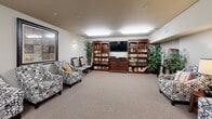 Located in Covington, WA, this single-floor amenity space spans a generously sized footprint and is organized into four distinct functional areas, each designed to support comfort and an active lifestyle. The lounge area features plush patterned armchairs, decorative framed artwork, and warm accent lighting, creating an inviting setting for relaxed conversation or quiet downtime. Neutral-toned carpet throughout the space contributes to a cohesive, comfortable atmosphere. An adjacent media and library room houses built-in dark wood bookcases stocked with books and media, flanking a wall-mounted flat-screen television. Tall potted plants add a natural element, while flush-mounted ceiling lights provide consistent, even illumination. A secondary seating alcove offers additional upholstered chairs arranged around a side lamp table, with framed art on the walls and direct access to a hallway corridor. The fitness room stands out with multiple windows on two walls that bring in natural light and outdoor views of the surrounding landscape. It is equipped with treadmills, stationary bikes, and exercise equipment, along with a large wall mirror that enhances the sense of space. Together, these areas form a well-rounded, community-focused facility.