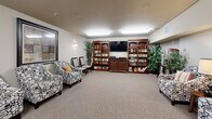 Located in Covington, WA, this single-floor amenity space spans a generously sized footprint and is organized into four distinct functional areas, each designed to support comfort and an active lifestyle. The lounge area features plush patterned armchairs, decorative framed artwork, and warm accent lighting, creating an inviting setting for relaxed conversation or quiet downtime. Neutral-toned carpet throughout the space contributes to a cohesive, comfortable atmosphere. An adjacent media and library room houses built-in dark wood bookcases stocked with books and media, flanking a wall-mounted flat-screen television. Tall potted plants add a natural element, while flush-mounted ceiling lights provide consistent, even illumination. A secondary seating alcove offers additional upholstered chairs arranged around a side lamp table, with framed art on the walls and direct access to a hallway corridor. The fitness room stands out with multiple windows on two walls that bring in natural light and outdoor views of the surrounding landscape. It is equipped with treadmills, stationary bikes, and exercise equipment, along with a large wall mirror that enhances the sense of space. Together, these areas form a well-rounded, community-focused facility.