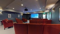 This private screening room is thoughtfully designed to deliver an immersive, cinema-quality experience. The space is anchored by a large wall-mounted projection screen at the front, paired with a ceiling-mounted projector that ensures a crisp, vivid picture. Flanking the screen, freestanding floor speakers provide robust audio coverage throughout the room. Seating consists of generously sized, deep red upholstered armchairs arranged in tiered rows, each equipped with built-in cup holders for added convenience. The layout accommodates a comfortable number of viewers while maintaining clear sightlines to the screen from every seat. The walls are finished in a rich slate blue-grey tone, enhancing the cinematic atmosphere and reducing light reflection. Warm sconce lighting along the side walls provides subtle ambient illumination, while recessed ceiling lights offer additional brightness when needed. Framed classic movie posters serve as tasteful decorative accents, reinforcing the room's dedicated entertainment purpose. Along the rear side wall, a built-in wet bar with dark cabinetry, a beverage refrigerator, and counter space adds a hospitality element ideal for entertaining. Charcoal drapery along the front wall frames the screen and contributes to the room's acoustics and light control. A door at the rear provides convenient access to an adjacent outdoor area.