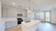 Located in Austin, TX, this well-appointed 2-bedroom, 2-bathroom single-story home offers a thoughtfully designed layout with modern finishes throughout. The open-concept kitchen serves as the heart of the home, featuring a large center island with a white quartz countertop and undermount sink, crisp gray shaker cabinetry, a subway tile backsplash, and a full suite of stainless steel appliances including a french door refrigerator, range, and built-in microwave. Stylish pendant lights with leather accents hang above the island, complementing the light wood-look flooring that flows through the main living areas. The living room receives ample natural light through large windows and offers access to an outdoor balcony area. Both bedrooms are carpeted for comfort, with the generously sized primary suite featuring an en-suite bathroom complete with a walk-in glass shower, marble-style tile flooring, a wide vanity with white countertop, and built-in shelving. The second bathroom includes a full tub-shower combination with white tile surround. Two closets provide organized storage, including a walk-in closet with built-in shelving and hanging rods. Clean white walls and recessed lighting throughout lend the home a bright, contemporary ambiance.