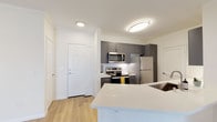 This single-floor residence presents a cohesive modern design with a neutral color palette and thoughtful layout. The open-concept kitchen serves as a central feature, showcasing charcoal gray cabinetry with sleek hardware, a white quartz peninsula countertop with an undermount sink, and a full suite of stainless steel appliances including a range, over-the-range microwave, and refrigerator. A white subway tile backsplash adds a clean, classic contrast to the darker cabinetry. Light wood-look flooring runs throughout the main living areas, complementing the bright white walls and trim.

The living area is generously proportioned, featuring crown molding, a ceiling fan, and a sliding glass door that opens to a balcony, allowing natural light to fill the space. The bedrooms are finished with plush carpeting, ceiling fans, and large windows fitted with horizontal blinds, providing a comfortable and well-lit atmosphere. One bedroom includes access to a walk-in or reach-in closet area.

The bathroom is cleanly appointed with a tub-shower combination, white tile surround, wood-look flooring, a gray vanity with white quartz countertop, and a large frameless mirror, maintaining the consistent modern aesthetic found throughout the home.