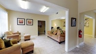 Located at 2414 Stone Rd in East Point, Georgia, this single-story community facility offers a well-organized and inviting layout designed for comfort and functionality. The interior is unified by light-toned ceramic tile flooring throughout and warm, neutral yellow-beige walls that create a bright and welcoming atmosphere.

The main lounge area is spacious and open, featuring vaulted ceilings that enhance the sense of volume. Comfortable upholstered seating, including a sofa and armchairs with accent pillows, is arranged for relaxed gathering. A sunlit sitting nook with large windows and vertical blinds provides views of the surrounding greenery, offering a quiet retreat within the facility.

A dedicated dining area accommodates multiple glass-top tables with white wicker chairs featuring floral cushions, suitable for group meals or meetings. Adjacent to this space is a fitness room, visible through an open doorway, equipped with cardio equipment including a treadmill.

The facility also includes clearly marked restroom facilities and additional rooms that appear to serve as office or resource spaces, with bulletin boards and informational materials visible. Framed artwork is displayed throughout the common areas, adding a polished, welcoming touch. Security is supported by mounted cameras. Natural light flows in generously from multiple windows, enhancing the overall openness of the space.