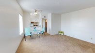This single-floor residence offers a practical and comfortable layout designed for everyday living. The main living area is generously sized, featuring neutral wall-to-wall carpeting that adds warmth underfoot. A ceiling fan is centrally positioned, providing both airflow and a subtle decorative touch. A textured ceiling adds dimension to the space, and a large window along the side wall invites ample natural light into the room.

The kitchen is open to the living area and features white cabinetry providing solid storage options, along with visible appliances and a functional counter workspace. The seamless visual connection between the kitchen and living space creates a cohesive, easy-to-navigate floor plan. A hallway leads from the main living area to the remaining rooms, suggesting a thoughtful separation between the living and private spaces.

The overall aesthetic is clean and neutral, with white walls and light-toned finishes that create a bright and airy atmosphere. The open arrangement of the living space allows for flexible furniture configurations, accommodating a range of lifestyles. This single-level layout is well-suited for those seeking a straightforward, accessible living environment with functional design elements throughout.