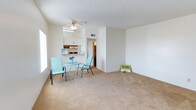 This single-floor residence offers a practical and comfortable layout designed for everyday living. The main living area is generously sized, featuring neutral wall-to-wall carpeting that adds warmth underfoot. A ceiling fan is centrally positioned, providing both airflow and a subtle decorative touch. A textured ceiling adds dimension to the space, and a large window along the side wall invites ample natural light into the room.

The kitchen is open to the living area and features white cabinetry providing solid storage options, along with visible appliances and a functional counter workspace. The seamless visual connection between the kitchen and living space creates a cohesive, easy-to-navigate floor plan. A hallway leads from the main living area to the remaining rooms, suggesting a thoughtful separation between the living and private spaces.

The overall aesthetic is clean and neutral, with white walls and light-toned finishes that create a bright and airy atmosphere. The open arrangement of the living space allows for flexible furniture configurations, accommodating a range of lifestyles. This single-level layout is well-suited for those seeking a straightforward, accessible living environment with functional design elements throughout.