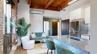 Located at 800 Broadway Blvd in the heart of Kansas City, MO, this one-bedroom, two-bathroom loft blends historic industrial character with modern finishes. Soaring ceilings with exposed wooden beams and visible ductwork give the open-concept living and kitchen area a striking urban aesthetic. Original hardwood floors run throughout, adding warmth and continuity to the space.

The kitchen is well-appointed with stainless steel appliances, white cabinetry, and a granite-topped island with bar seating — ideal for casual dining or entertaining. A dramatic exposed brick accent wall anchors the kitchen and living area, complementing the industrial design language. Large black-trimmed windows flood the space with natural light and offer views of the surrounding downtown streetscape.

The bedroom is a comfortable retreat, featuring the same soaring ceilings, original hardwood floors, and generous natural light from dual windows with city views. Both bathrooms are cleanly finished with white vanities, modern fixtures, and hardwood flooring, maintaining a consistent design palette throughout. A hallway provides practical connectivity between rooms. This loft captures the character of a historic Kansas City building reimagined for contemporary urban living.