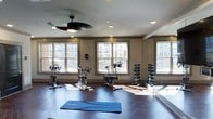 Located at 811 Spring Forest Rd in Raleigh, this fitness center offers a bright and functional workout environment designed to support a variety of exercise routines. The space features rich hardwood-style flooring that adds warmth and durability underfoot, complemented by neutral-toned walls that create a clean, focused atmosphere. Large windows line the walls, flooding the interior with natural light and providing views of the surrounding building exterior, enhancing the sense of openness throughout the room. Recessed lighting and a ceiling fan with an integrated light fixture provide additional illumination and climate comfort. The workout area is equipped with multiple stationary cycling bikes arranged to take advantage of the natural light, making it well-suited for cardio training. A tiered dumbbell rack stocked with free weights is positioned near a floor-to-ceiling mirror along one wall, supporting strength training exercises. A yoga or exercise mat on the floor indicates space for stretching and floor-based workouts. The mirrored wall adds depth to the room while allowing exercisers to monitor their form. Overall, the space presents a well-organized, inviting fitness environment.