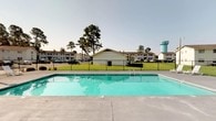 This single-story residential community offers a welcoming outdoor environment centered around a generously sized swimming pool with clear, turquoise water. The pool area is finished with a wide concrete surround, providing ample space for lounging, and is enclosed by a black metal fence for safety and definition. Steps with a metal handrail offer easy access into the water.

The grounds are well-maintained, featuring neatly trimmed lawns that extend throughout the community. Tall, mature pine trees punctuate the landscape, lending a sense of natural character and shade to the property. Multiple residential buildings are visible in the background, constructed in a classic style with white and neutral-toned exteriors and pitched roofs, contributing to a cohesive community aesthetic.

A dedicated parking area is accessible from the grounds, accommodating residents with convenient off-street parking. A distinctive water tower is visible on the horizon, adding a notable landmark to the skyline. The overall ambiance of the community is open and airy, with expansive views of the surrounding landscape under a clear, open sky — making the outdoor spaces a central and inviting feature of daily life here.