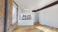 Located at 1011 N. Hancock St. in Philadelphia, PA, this loft-style unit at Carriage Wheel Lofts seamlessly merges industrial heritage with contemporary design. The open-concept living and kitchen area anchors the space, featuring light hardwood flooring that extends throughout, crisp white walls, and exposed original brick that lends unmistakable character. A striking industrial ductwork system runs overhead, reinforcing the loft aesthetic.

The kitchen is thoughtfully designed with a large white peninsula with a quartz countertop, stainless steel appliances including a full-size refrigerator and microwave, and a classic white subway tile backsplash. White cabinetry keeps the space feeling bright and clean, while a window brings in natural light.

The bedroom areas showcase dramatic exposed brick walls spanning multiple windows that flood the rooms with natural light. The industrial ductwork continues here, complementing the raw architectural details and creating a cohesive loft atmosphere.

A well-appointed bathroom features large-format neutral tiles, a glass-enclosed walk-in shower, and a sleek white vanity. A clean, light-filled hallway with recessed lighting connects the various spaces, also providing closet storage. Throughout, the interplay of original brick, modern finishes, and abundant natural light creates a distinctly urban yet refined living environment.