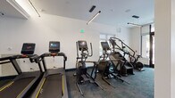 This well-appointed fitness center in Savannah, GA offers a thoughtfully designed, single-floor layout divided into distinct workout zones. The cardio area is equipped with multiple treadmills, elliptical trainers, and stair climbers, all featuring digital displays. Ceiling fans and linear LED track lighting ensure a comfortable and well-lit environment, while large windows invite abundant natural light and outdoor views of the surrounding streetscape.

The strength training zone features a rack system in bold red accents stocked with medicine balls and resistance equipment, a full dumbbell rack, an adjustable bench, and wall-mounted flat-screen TVs for entertainment during workouts. Mirrored walls amplify the sense of space and allow users to monitor their form. A ceiling fan overhead adds to the room's functionality.

A separate, open studio area provides a bright, airy space ideal for yoga, stretching, or group classes. This room boasts floor-to-ceiling windows, light tile flooring, a decorative chandelier, and yoga mats for use. A striking navy laser-cut decorative panel serves as a sophisticated room divider, adding an elegant architectural accent that distinguishes this fitness center's refined aesthetic. Throughout, the clean white walls, dark teal accents, and quality flooring create a polished and motivating atmosphere.
