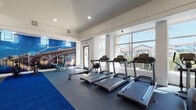 This fitness center is a spacious, single-floor facility designed to accommodate a wide range of workout routines. The layout is open and generously proportioned, with elevated ceilings and recessed lighting that create a clean, energizing atmosphere throughout the space.

Large floor-to-ceiling windows line multiple walls, allowing abundant natural light to fill the room and offering views of the surrounding outdoor area, including what appears to be a pool and residential buildings beyond. Two exterior doors provide convenient direct access to the outdoor amenity areas.

The flooring consists of durable rubber gym material in a dark speckled finish, complemented by a bold blue carpet runner that bisects the central workout area, providing a dedicated space for functional training and floor exercises.

Cardio equipment is prominently featured along the windowed walls and includes multiple treadmills, an elliptical trainer, a stationary bike, and a rowing machine. The strength training zone is well-stocked with a full rack of dumbbells, adjustable benches, a power rack with weight plates, a cable machine, and a sled for functional training.

A wall-mounted television is positioned centrally for visibility across the floor. A striking large-format photographic mural depicting a vibrant nighttime cityscape with a lit bridge serves as a bold focal point, adding visual energy to the space. Full-length mirrors on one wall enhance the sense of depth and functionality.
