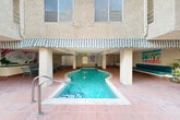 This courtyard amenity space is centrally located within a multi-story stucco building complex, offering residents a well-designed outdoor retreat. At the heart of the space sits a freeform swimming pool with a classic kidney-inspired shape and striking turquoise water, complemented by a separate spa with step-in access and decorative tile detailing. The pool and spa are framed by a spacious terracotta-tiled deck, providing ample room for lounging and movement.

A covered lounge area, sheltered by a distinctive corrugated green-trimmed awning supported by columns, extends along one side of the pool. This shaded zone features built-in seating with teal cushions, textured stucco walls, and colorful artwork that adds a lively, artistic character to the space. Glass block wall accents and recessed lighting contribute to the functional design of the covered area, which also includes a dining table and chairs for outdoor gatherings.

The open courtyard portion is generously proportioned, with terracotta tile extending throughout and lined with an array of potted trees, hedging, and greenery that provide privacy and a sense of lush enclosure. The surrounding multi-story building facades, featuring balconies and louvered windows, frame the space on multiple sides, creating an intimate, resort-like atmosphere within the complex.