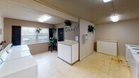 Located at 2627 Wimpole Ave in Knoxville, Tennessee, this single-floor shared laundry facility is designed to provide a functional and practical space for residents. The layout is open and well-organized, featuring multiple commercial-grade top-loading washing machines and dryers lined along the walls, offering ample capacity for laundry needs. Dedicated folding counters are positioned throughout the space, providing convenient surfaces for sorting and folding clothes. The flooring consists of light-colored vinyl tile, and the textured ceilings are fitted with fluorescent lighting that ensures bright, even illumination throughout. Exposed copper piping runs along the upper walls, adding an industrial character to the space. The facility includes a windowed exterior door that allows for natural light and ventilation, complemented by a window at the front of the room. Decorative touches such as potted greenery and framed artwork on the walls contribute to a welcoming atmosphere. Informational notices are posted for user convenience. The space is clearly marked with laundry signage, making it easy to navigate. Overall, this laundry facility is a well-equipped, communal utility space designed with both efficiency and practicality in mind.