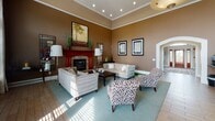 This single-floor space is defined by its dramatic, soaring ceilings accented with crisp crown molding and warm earth-toned walls that create an inviting and sophisticated atmosphere throughout. The main lounge area is a standout feature, centered around a striking stone-surround fireplace with a rich cherry wood mantel. Plush neutral sofas, patterned accent chairs, and a soft area rug are arranged to encourage conversation, while recessed lighting and generous natural light streaming through tall, multi-pane windows with elegant drapery complete the welcoming ambiance. Adjacent to the lounge, a casual bistro-style seating area offers high-top round tables and upholstered bar stools positioned near a wall of floor-to-ceiling windows, providing an abundance of natural light. A substantial built-in cherry wood entertainment wall with display shelving and a flat-screen television anchors this zone, adding both function and visual warmth. The kitchen is appointed with rich cherry cabinetry, dark countertops, and a wraparound bar-height peninsula that comfortably accommodates seating for multiple guests. Full-sized appliances and ample storage make this space highly functional. Light wood-look flooring flows consistently throughout, tying each area together into a cohesive and polished whole.