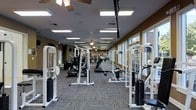 This fitness center offers a generously proportioned, single-floor workout space designed to accommodate a comprehensive range of exercise needs. The room is laid out with an elongated floor plan, creating a sense of depth and openness as equipment lines extend from front to back. A row of large windows along one side of the space floods the interior with natural light, providing views of the outdoor surroundings and enhancing the overall atmosphere. The ceiling features recessed panel lighting supplemented by ceiling fans, ensuring consistent illumination and air circulation throughout. The floor is covered in a durable, multi-toned carpet designed to withstand heavy use while providing some cushioning underfoot. Strength training is well represented, with multiple cable machines, weight stack units, and resistance equipment arranged in an organized fashion along both sides of the room. Cardio machines, including treadmills and ellipticals, are positioned to take advantage of the natural light from the windows. The neutral color palette of tan walls and gray flooring contributes to a clean, focused environment suitable for a variety of fitness routines.