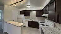 This two-story residence offers a practical and well-appointed layout across two levels. The main floor welcomes visitors with a carpeted living area bathed in natural light from a large front-facing window, with a staircase featuring sleek black metal railings leading to the upper level. The kitchen is a standout feature, showcasing dark espresso-toned cabinetry with raised-panel detailing, light granite countertops, a classic subway tile backsplash, and white appliances including a refrigerator, dishwasher, and electric range. Track lighting and a flush-mount ceiling fixture provide ample illumination, while tile flooring adds a clean, durable finish to this functional space. The bathroom is finished with large-format gray wall tiles surrounding a soaking tub and shower combination, complemented by mosaic tile flooring and a vanity topped with granite-patterned counters and dark cabinetry below. The bedrooms are carpeted and neutral in tone, offering versatile spaces with textured walls, panel-style doors, and ceiling ventilation. Throughout the home, textured ceilings, neutral paint tones, and consistent trim details contribute to a cohesive and move-in ready aesthetic.