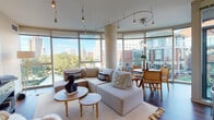 Located at 2 Center St in the heart of Newark, NJ, this single-floor residence offers a thoughtfully designed layout with modern finishes throughout. The open-concept living and dining area is a standout feature, wrapped in sweeping curved floor-to-ceiling windows that flood the space with natural light and frame striking views of the Newark skyline and surrounding greenery. Track lighting runs along the ceiling, adding both functionality and a contemporary aesthetic to the space. Rich hardwood flooring anchors the living area and transitions seamlessly into the kitchen, which is outfitted with white cabinetry, dark stone countertops, and stainless steel appliances, including a full-size refrigerator. A built-in desk area provides a dedicated workspace. The primary bedroom is generously proportioned, featuring large windows with tree-lined views, plush carpeting, and a serene, neutral palette. A second bedroom also benefits from abundant natural light. Two well-finished bathrooms serve the residence — one featuring a glass-enclosed walk-in shower with large-format tile, and the other a soaking tub, both with white cabinetry and dark granite countertops. The overall ambiance is clean, contemporary, and urban.