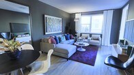 This single-floor space showcases a well-appointed living area that blends contemporary design with vibrant personality. The room is anchored by deep charcoal-gray walls, which provide a sophisticated backdrop for a large, colorful abstract artwork — a striking focal point that draws the eye immediately upon entry. Light hardwood flooring runs throughout, offering a warm contrast to the cooler wall tones.

The living area is generously proportioned, centered around a plush gray sectional sofa adorned with an eclectic mix of decorative pillows in jewel tones — cobalt blue, teal, and purple — adding layers of color and texture. A deep purple area rug grounds the seating arrangement, complementing the bold design choices throughout. A sleek white tulip-style coffee table sits at the center, paired with two clean-lined white armchairs that introduce a modern, minimalist balance.

Natural light filters generously through a large window dressed with sheer white curtains, offering views of the surrounding exterior. A tall arc floor lamp provides additional ambient lighting. Near the window, a globe and sculptural accents lend an artful, curated quality to the space, reflecting a confident and cohesive interior aesthetic.