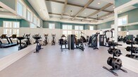 Located in Santa Rosa Beach, Florida, this fitness center offers a generously sized, single-floor exercise room designed to support a full range of workout routines. The space is immediately distinguished by its soaring coffered ceilings accented with exposed wooden beams and recessed lighting, creating an open, airy atmosphere that feels both functional and inviting.

The walls are finished in a calming teal hue above white wainscoting, lending the space a clean, coastal aesthetic consistent with its Florida Panhandle setting. Large multi-pane windows line the walls on multiple sides, flooding the interior with natural light and offering views of the surrounding outdoor environment. Full-length mirrors along one wall further enhance the sense of space and provide practical utility for form-checking during workouts.

The exercise room is comprehensively equipped with a wide selection of strength and cardio machinery. Strength equipment includes Hoist power racks, free weight benches, a tiered dumbbell rack, and multiple Spirit-brand cable and resistance machines. The cardio section features recumbent bikes, upright cycles, elliptical trainers, and rowing machines. Ceiling fans mounted from the coffered ceiling provide additional air circulation throughout the space.

French doors provide a welcoming entry point, and the durable, light-toned flooring throughout supports heavy equipment while maintaining a polished appearance. This fitness center is a well-appointed, purpose-built facility suited for serious and casual exercisers alike.
