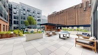 Located at 108 Hamilton St in Bound Brook, NJ, this thoughtfully designed outdoor amenity space serves as a central gathering hub set within a multi-story residential community. The courtyard blends modern design with industrial character, drawing on the surrounding brick and gray panel architecture to create a cohesive urban aesthetic.

The space is organized into several distinct zones catering to a range of activities. A lush lawn area features Adirondack chairs arranged around a fire pit, providing a relaxed setting for socializing beneath the open sky. Adjacent to this, a spacious tiled patio accommodates outdoor dining furniture and lounge seating, framed by manicured planters and native landscaping.

A standout feature is the covered pavilion, which houses a fully outfitted outdoor kitchen complete with stainless steel grills, white quartz countertops, brick base cabinetry, and a dishwasher — all sheltered beneath a sleek pergola structure with a wood-paneled ceiling and ceiling fans. A dedicated sunbathing terrace with teak chaise lounges and a navy accent wall with decorative panels adds a resort-like touch.

The grounds are enriched by raised planters, accent lighting, and a ping-pong table, making this courtyard a well-rounded outdoor retreat for residents of the community.