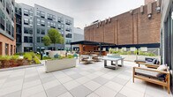Located at 108 Hamilton St in Bound Brook, NJ, this thoughtfully designed outdoor amenity space serves as a central gathering hub set within a multi-story residential community. The courtyard blends modern design with industrial character, drawing on the surrounding brick and gray panel architecture to create a cohesive urban aesthetic.

The space is organized into several distinct zones catering to a range of activities. A lush lawn area features Adirondack chairs arranged around a fire pit, providing a relaxed setting for socializing beneath the open sky. Adjacent to this, a spacious tiled patio accommodates outdoor dining furniture and lounge seating, framed by manicured planters and native landscaping.

A standout feature is the covered pavilion, which houses a fully outfitted outdoor kitchen complete with stainless steel grills, white quartz countertops, brick base cabinetry, and a dishwasher — all sheltered beneath a sleek pergola structure with a wood-paneled ceiling and ceiling fans. A dedicated sunbathing terrace with teak chaise lounges and a navy accent wall with decorative panels adds a resort-like touch.

The grounds are enriched by raised planters, accent lighting, and a ping-pong table, making this courtyard a well-rounded outdoor retreat for residents of the community.