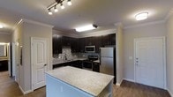 This single-floor residence showcases a thoughtfully designed interior with a strong emphasis on modern style and functional living. The kitchen serves as the heart of the space, outfitted with rich, dark espresso cabinetry that extends to the ceiling and is complemented by a decorative mosaic tile backsplash in a contrasting black-and-white pattern. Stainless steel appliances, including a refrigerator, electric range, built-in microwave, and dishwasher, lend a sleek, contemporary feel. A central island with a light granite countertop provides generous prep space and casual seating, making it an ideal gathering spot. Crown molding runs along the ceiling throughout, adding a refined architectural touch, while durable hardwood-style flooring grounds the space with warmth. Track lighting and a flush-mount ceiling fixture ensure the kitchen is well-lit for both cooking and entertaining. The neutral wall tones offer a versatile backdrop that complements the bold cabinetry. Doorways visible from the kitchen hint at additional rooms beyond, suggesting a practical and connected floor plan. Overall, the space balances classic detailing with modern finishes for a polished, inviting atmosphere.