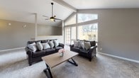 This single-floor space is defined by its striking vaulted ceilings with exposed beam accents, creating an airy, expansive atmosphere throughout. The open-concept layout seamlessly connects multiple functional zones, beginning with a generously sized living area anchored by two dark leather sofas arranged around a wooden X-frame coffee table. Ceiling fans are positioned throughout, enhancing both comfort and the architectural character of the high ceilings.

A tile-surround corner fireplace serves as a focal point between the living and dining areas, flanked by decorative wall art and tall accent pieces. Adjacent to the fireplace, a round dining table with dark leather chairs provides an inviting space for gatherings. A second dining area near the kitchenette offers additional seating options.

The kitchenette features warm wood cabinetry, counter space, and built-in appliances, making the space well-equipped for entertaining. Large windows throughout flood the interior with natural light and offer views of the surrounding landscape. Plush neutral carpeting runs throughout, complementing the warm gray walls and contributing to the overall cohesive, welcoming ambiance of the space.
