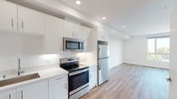 This thoughtfully designed one-bedroom, one-bathroom unit offers a clean, modern aesthetic with consistent warm-toned hardwood flooring and crisp white walls flowing throughout the entire space. The open-concept kitchen and living area serves as the heart of the unit, featuring sleek white flat-panel cabinetry that extends to the ceiling for maximum storage, white quartz countertops, a stainless steel appliance suite including a refrigerator, electric range, over-the-range microwave, and dishwasher, and recessed lighting that brightens the space. The kitchen transitions seamlessly into the living area, creating a versatile and airy layout. The bedroom is a comfortable, well-proportioned room bathed in natural light from a large window that frames views of palm trees and the surrounding neighborhood. The bedroom connects to a generously sized walk-in closet outfitted with built-in wire shelving systems along multiple walls, offering excellent organization and storage capacity. The full bathroom features a dark wood-tone vanity with white countertop and an undermount sink, a large mirror, and hardwood-style flooring that ties the room cohesively to the rest of the unit. The overall ambiance is bright, clean, and contemporary.