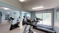 This fitness center offers a clean, functional environment designed to support a variety of cardio workouts. The room is finished with soft blue walls and white crown molding, creating a calm and inviting atmosphere. Decorative white columns add a subtle architectural detail that divides the space while maintaining an open feel. The flooring features a speckled, durable surface well-suited for an active environment. A suite of Spirit-brand cardio equipment is the centerpiece of the space, including treadmills, an elliptical trainer, and stationary bikes — providing users with a comprehensive range of aerobic exercise options. Large windows fitted with vertical blinds line one wall, flooding the room with natural light and offering pleasant views of the lush greenery outside. Overhead fluorescent lighting ensures the space remains well-lit at all times. A wall clock is mounted near the mirrored section, allowing users to track their workout time. The mirrors along one wall enhance the sense of space and allow users to monitor their form. Overall, this fitness center balances practicality with a welcoming aesthetic, making it a comfortable and effective space for daily exercise.