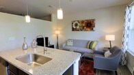 This single-floor space showcases an open-concept layout that connects the kitchen and living area in a cohesive, functional design. The kitchen features a standout granite countertop with a speckled finish, housing a stainless steel undermount sink equipped with a sleek, high-arc pull-down faucet. A pendant light fixture hangs above, casting warm, focused light over the kitchen workspace and adding a modern touch to the overall design.

The transition into the living area is smooth and unobstructed, creating a sense of openness throughout the space. The living area is anchored by a contemporary gray sofa accented with colorful throw pillows in green, yellow, and patterned designs, paired with a matching chair. A rich, deep-red area rug adds warmth and contrast against the hardwood-style flooring. A floor lamp provides ambient lighting alongside the pendant fixtures, while a flat-screen television is positioned on a low-profile media console against the wall. Decorative wall art featuring bold floral motifs adds a vibrant focal point to the space. The overall ambiance is modern, welcoming, and thoughtfully curated.