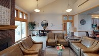 This thoughtfully designed single-floor space offers a warm and inviting atmosphere, highlighted by consistent warm wood trim, vaulted ceilings, and large windows that flood each area with natural light. The main lounge area features soaring ceilings with elegant pendant lighting, comfortable leather seating arranged around a central coffee table, and a decorative console table with a circular mirror accent. A secondary gathering room showcases a full brick fireplace flanked by built-in shelving with media storage, a flat-screen television, and direct access to an outdoor patio area through glass doors, extending the living space further. A dedicated dining area is lined with a generous bank of wood-framed windows offering views of the surrounding landscape, creating a bright and airy setting around a solid wood dining table. The fully equipped kitchen features rich wood cabinetry, tile flooring, light stone countertops, a double sink, and black appliances, complemented by another row of large windows that maintain the space's connection to the outdoors. Throughout, neutral wall tones, carpet flooring in living areas, and cohesive warm wood detailing create a unified, comfortable, and functional environment.