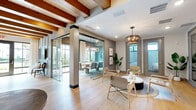Located at 7950 Park Blvd N in Pinellas Park, Florida, this thoughtfully designed single-floor space blends coastal charm with modern functionality. Light hardwood flooring flows seamlessly throughout, complemented by crisp white walls and an abundance of natural light streaming in through large steel-framed windows and glass doors that frame lush outdoor views.

The space features multiple dining and conference rooms, each with its own distinct character. A standout conference room showcases floor-to-ceiling steel-framed windows dressed with elegant draping curtains, a solid wood communal table surrounded by contemporary chairs, a wall-mounted display screen, and a geometric pendant light fixture overhead — all set against a muted blue-grey accent wall with a shiplap ceiling detail.

A separate office or meeting area benefits from dramatic exposed wood ceiling beams, direct outdoor access through glass-paned French doors, a built-in refreshment station with cabinetry, and ample natural light that gives the room an airy, open feel.

The central living area serves as a welcoming hub, featuring a woven pendant light fixture, a round marble-topped coffee table, and modern lounge chairs arranged on a natural jute rug. Glass partitions elegantly divide the spaces without sacrificing openness. A bathroom completes the layout, making this a fully equipped, professionally styled facility.