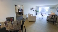 This single-floor space is thoughtfully laid out to maximize functionality and comfort. The living area serves as the central hub, featuring neutral-toned carpeting and a plush cream sofa positioned to take full advantage of a large sliding glass door that floods the room with natural light and provides access to an outdoor balcony or patio. Wall-mounted shelving displays tasteful decorative accents, adding character to the otherwise clean, white walls. A potted plant near the sliding door brings a touch of greenery to the space. Adjacent to the living area, a dining space is anchored by a striking dark marble-topped table paired with dark wood chairs, offering a bold contrast to the lighter surroundings. Through an open doorway, a dedicated office or study room is visible, outfitted with a desk, task lighting, and seating — ideal for productive work or focused tasks. The carpeted flooring throughout the main living areas contributes to a warm, comfortable ambiance, while baseboard heating ensures year-round comfort. Overall, the space presents a practical and well-organized layout with a calm, neutral aesthetic.