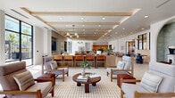 This expansive single-floor amenity space offers a thoughtfully designed environment that blends contemporary architecture with warm, natural materials. The open-plan layout is anchored by a spacious central lounge area featuring structured wood-framed seating, woven area rugs, and a distinctive coffered ceiling with exposed light wood beams and recessed lighting that creates a refined yet welcoming atmosphere.

Large floor-to-ceiling windows and glass wall systems line multiple sides of the space, flooding the interiors with natural light and offering views of the surrounding outdoor pool deck and landscape. Light-toned large-format tile flooring runs throughout, enhancing the sense of openness and continuity.

A built-in wood shelving unit serves as a central display and workspace feature, while a dedicated library corridor lined with illuminated built-in bookshelves and a patterned runner rug adds an intimate, functional touch. Interior glass windows between rooms provide visual connectivity while maintaining defined zones.

The game room stands out with bold teal accent walls, oversized black-framed windows showcasing hillside views, a pool table, and a long communal bar-height island — ideal for social gatherings. A separate area features a shuffleboard table and cozy reading nook with built-in shelving. Arched architectural details and private rooms with glass-panel doors round out the diverse, amenity-rich offering of this well-appointed space.
