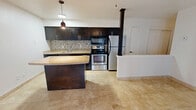 This efficiently designed single-floor unit makes excellent use of its compact footprint, offering an open-concept layout that seamlessly combines the kitchen, dining, and living areas into one cohesive space. The kitchen is a standout feature, outfitted with rich dark-wood cabinetry, granite-style countertops, a mosaic tile backsplash, and a full suite of stainless steel appliances including a range, microwave, refrigerator, and dishwasher. A peninsula with a matching countertop provides additional prep space and serves as a natural divider between the kitchen and living area. Pendant lighting above the peninsula adds a warm, functional accent. The living area is bright and open, benefiting from a glass-paneled exterior door and an adjacent window that invite natural light, complemented by a ceiling fan overhead. Consistent light-toned stone tile flooring runs throughout the entire unit, lending a clean and cohesive aesthetic. The full bathroom features a tub-shower combination with white tile surround, tile flooring, and a granite-topped vanity. Two closets provide practical storage, and a hallway connects the spaces efficiently. The overall ambiance is clean, functional, and modern, making this a practical and attractive space.