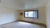 This single-floor space offers a clean, functional layout designed for comfortable living. The main room is characterized by its light-colored walls and smooth ceiling, which together create a bright and airy atmosphere. A large, wood-framed picture window serves as a central architectural feature, allowing generous amounts of natural light to fill the room while offering views of the surrounding greenery outside. Sheer curtains provide a soft, diffused quality to the incoming light. The room is fitted with wall-to-wall carpeting in a neutral tone, lending a warm and quiet underfoot feel throughout the space. Climate control is well-considered, with a baseboard heating system running along the exterior wall beneath the window, ensuring consistent warmth during cooler months. A wall-mounted air conditioning unit provides cooling capability during warmer seasons, making the space comfortable year-round. The overall ambiance is simple and understated, with a neutral color palette that provides a versatile backdrop suitable for a variety of uses. The space is straightforward in its design, prioritizing functionality and practicality while maintaining a tidy, livable environment.