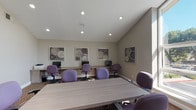 Located at 1700 Via Pacifica in Corona, California, this single-floor community space is thoughtfully designed to serve as both a functional business center and a comfortable gathering lounge. The layout flows seamlessly between a dedicated computer workstation area and an inviting lounge space, connected by glass-paneled French doors that lead to an adjoining room with a kitchen and bar seating.

The business center section features a built-in wood-tone desk along the wall, outfitted with multiple computer workstations and a printer. Purple upholstered task chairs surround both the workstations and a central conference table, creating a cohesive and productive workspace. A series of framed black-and-white botanical artworks lines the walls, adding a polished, decorative touch throughout.

The lounge area transitions naturally from the business center, offering a neutral-toned sofa accented with purple and white throw pillows, a side table with a lamp, a decorative mirror, and lush indoor plants. Soaring vaulted ceilings with recessed lighting and large windows and glass doors flood the space with natural light and provide views of the surrounding outdoor area.

The patterned carpet, neutral wall tones, and consistent purple accent palette tie the entire space together, creating a professional yet welcoming environment suited for both focused work and casual relaxation.