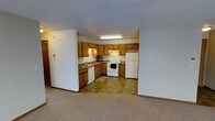 This single-floor residential space offers a practical and functional layout designed for comfortable everyday living. The kitchen is a standout feature, showcasing warm honey-toned wood cabinetry that lines the walls in a classic L-shaped configuration, providing generous storage and counter space. The kitchen is fully equipped with a white appliance suite, including an electric range, dishwasher, and a full-sized refrigerator, making it move-in ready. A built-in sink is positioned along the counter, and a flush-mounted fluorescent light fixture ensures bright, even illumination throughout the cooking area. The flooring in the kitchen features durable stone-look tile, offering both style and easy maintenance. The kitchen flows seamlessly into the adjacent living area, which is finished with plush wall-to-wall carpeting that adds warmth and comfort underfoot. The neutral color palette throughout — from the white walls and ceilings to the light carpet — creates a clean, bright, and welcoming ambiance. The open sightlines between the kitchen and living spaces make the overall floor plan feel connected and spacious, ideal for both everyday routines and entertaining.