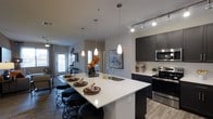 This single-floor residence showcases a thoughtfully designed open-concept layout that seamlessly connects the kitchen, dining, and living areas into one cohesive living space. The kitchen is a standout feature, anchored by a generous white quartz island with bar seating for multiple guests — ideal for casual dining or entertaining. Sleek dark cabinetry provides a striking contrast against the light countertops, while a classic subway tile backsplash adds texture and polish. Stainless steel appliances, including a range and over-the-range microwave, complete the modern culinary setup. A deep stainless steel sink sits beneath an elegant arc faucet, and the island is illuminated by a trio of teardrop pendant lights that add warmth and sophistication. Track lighting runs along the ceiling, offering flexible illumination throughout the space. Wide-plank light wood flooring flows throughout, enhancing the airy, contemporary feel. The adjoining living area is bathed in natural light from large windows, creating a bright and welcoming atmosphere. Neutral wall tones and modern finishes throughout contribute to a clean, cohesive aesthetic that is both functional and visually inviting.