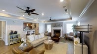 This single-floor residence offers a thoughtfully designed layout that balances comfort and functionality. The living room serves as the heart of the home, anchored by a classic white-mantel fireplace and framed by neutral gray walls that create a calm, inviting atmosphere. A coffered tray ceiling with dual ceiling fans adds architectural character and enhances the sense of spaciousness overhead, while recessed lighting provides warm, even illumination throughout.

The living area is generously proportioned, offering ample space for multiple seating arrangements. Decorative star-shaped wall accents above the fireplace add a personal touch, contributing to the room's relaxed, curated aesthetic. Natural light filters in through several windows, brightening the space and connecting the interior to the outdoors.

Adjacent to the living room, a bar-style kitchen is partially open to the main living space, featuring dark wood cabinetry, pendant lighting over a raised counter, and bar stool seating — a practical and social setup well-suited for casual dining and entertaining. The transition between spaces flows naturally, making the overall layout feel cohesive and easy to navigate. Neutral flooring ties the rooms together, reinforcing the home's warm and welcoming character throughout.