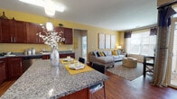 The Alterra at 647 Brook Street in Rocky Hill, Connecticut presents a thoughtfully designed 2-bedroom, 2-bathroom residence that combines modern finishes with a warm, inviting atmosphere. The open-concept kitchen, dining, and living area forms the heart of the home, anchored by a granite-topped island with bar seating. Rich cherry wood cabinetry, stainless steel appliances—including a gas range, microwave, and dishwasher—and hardwood-style flooring throughout give the kitchen a polished, functional appeal. The living room is bathed in natural light from large windows dressed with layered curtains, and features warm yellow accent walls that create an energetic yet comfortable ambiance. A dedicated dining area with hardwood flooring and deeper accent walls provides a refined space for entertaining. Both bedrooms are carpeted for comfort, each benefiting from natural light and generous closet space. The primary bedroom includes a walk-in closet, while both rooms offer a tranquil retreat feel. The primary bathroom features tile flooring and a glass-enclosed shower, while the second bathroom includes a tub-shower combination and a matching cherry wood vanity. Throughout the residence, consistent finishes and warm tones create a cohesive, modern living experience.