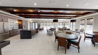 Located at 3011 Mesa Verde Dr in Orlando, Florida, this single-story community clubhouse is thoughtfully designed to offer a variety of social and recreational amenities within one cohesive space. The interior is unified by large-format light tile flooring, recessed lighting, and an abundance of natural light streaming through numerous windows dressed with woven wood shades.

The main gathering area is defined by dramatic dark wood ceiling beams paired with wood-paneled ceiling insets, lending warmth and architectural character to the open layout. Stylish pendant lighting illuminates multiple seating arrangements, including a generous lounge area with upholstered sofas and accent chairs, as well as several round and rectangular dining tables with rattan-style seating — all oriented toward views of an outdoor pool area through glass doors.

Adjacent to the lounge, a fully equipped kitchen features sleek dark cabinetry, stainless steel appliances including a full-size refrigerator and wall oven, a patterned tile backsplash, and a dark countertop island with bar seating. A dedicated game room nearby accommodates a full-size billiards table beneath a tray ceiling with exposed beams, complemented by natural light.

A separate, dedicated screening room rounds out the amenities, featuring tiered stadium-style seating, plush gray upholstered chairs with cup holders, wall sconces, carpeted floors, and a large mounted display screen — delivering a true theater experience.