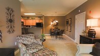 This single-floor residence offers an open-concept living arrangement that seamlessly connects the main living, dining, and kitchen areas into one cohesive and functional space. The interior is finished in warm, earthy tones — deep taupe walls paired with light neutral flooring create a grounded, inviting ambiance throughout.

The living area provides a comfortable gathering space, furnished with patterned upholstered seating that adds character and a sense of coziness. Crown molding runs along the ceiling, lending a refined architectural detail that elevates the overall finish of the space.

The dining area is positioned at the heart of the open floor plan, anchored by an elegant chandelier that casts a warm glow over the dining table and chairs. A decorative mirror and wall art further enhance the area's welcoming character.

The kitchen is positioned just beyond the dining area and features warm wood cabinetry, black appliances, and a raised breakfast bar that provides both additional workspace and a natural visual divider between the kitchen and the living spaces. Recessed and flush-mount lighting ensures the kitchen remains bright and functional.

Overall, the space presents a well-balanced blend of comfort and style, with consistent finishes and warm lighting that contribute to a relaxed yet polished atmosphere throughout.
