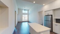 This single-floor residence showcases a contemporary design with an emphasis on height and natural light throughout. The open-concept living and dining area is defined by dramatically tall ceilings, dark hardwood-style flooring, and a ceiling fan overhead. A large window at the far end of the living space, positioned beneath a clerestory window, floods the area with abundant natural light and offers views of the surrounding neighborhood.

The kitchen is well-appointed with crisp white cabinetry, quartz countertops, and a bold tile backsplash. A central island provides additional prep and seating space, while stainless steel appliances—including a refrigerator, range, microwave, and dishwasher—complete the functional layout.

The bedrooms feature plush carpet flooring and soaring ceilings with generous window placements that invite natural light. Storage is well-addressed with both reach-in closets featuring wire shelving and a dedicated walk-in closet equipped with multi-tier shelving systems.

The bathroom is finished with light tile flooring, a wide vanity with a white countertop, a framed mirror, warm wall-mounted lighting, and a frosted clerestory window that maintains privacy while allowing natural light to enter.