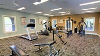 This fitness center offers a practical and comfortable environment for a full workout experience. The room features a dual-tone color scheme, with soft sage green and warm mustard yellow walls accented by crisp white trim, creating an inviting atmosphere. Multiple ceiling fans ensure consistent air circulation throughout the space, complementing the bright overhead fluorescent lighting. Natural light filters in through several windows positioned along the walls, adding to the room's openness.

The facility is equipped with a variety of cardio machines, including a treadmill and an elliptical trainer, providing options for cardiovascular exercise. A multi-station cable machine supports a range of strength training exercises, while a dedicated free weights area features a fully stocked dumbbell rack. A workout bench adds further versatility for strength training routines. Instructional fitness posters are displayed on the walls, serving as useful references during workouts. The floor is covered in durable, patterned carpet designed to withstand heavy equipment and frequent use. Overall, the space is thoughtfully arranged to maximize functionality while maintaining a clean and organized appearance.