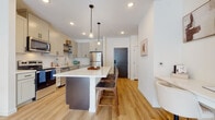 This thoughtfully designed single-floor residence offers a seamless blend of modern finishes and functional living spaces. The open-concept kitchen serves as the heart of the home, featuring soft gray shaker-style cabinetry, a white subway tile backsplash, stainless steel appliances including a refrigerator, range, microwave, and dishwasher, and a generous island with a crisp white countertop and bar seating for two. Warm-toned hardwood flooring flows throughout the main living areas, complemented by recessed lighting and pendant fixtures that create an inviting ambiance. The living room is bathed in natural light from a large sliding glass door that opens to an outdoor patio, offering views of the surrounding neighborhood. The bedroom is a serene retreat, carpeted for comfort, with oversized windows that bring in abundant daylight. It connects directly to a spacious walk-in closet with built-in shelving and a full bathroom. The bathroom is finished with light gray cabinetry, a white quartz vanity, a tub-shower combination, and warm hardwood-style flooring accented by matte black hardware throughout. Clean white walls and a neutral palette unify the space, creating a bright and contemporary atmosphere from every angle.