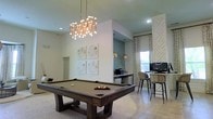 This single-floor amenity lounge is thoughtfully designed to blend recreation and relaxation in a sophisticated setting. The centerpiece of the space is a full-size billiards table in rich dark wood, positioned beneath a striking modern chandelier composed of exposed brass tubing and globe bulbs that serves as a dramatic focal point. Soaring ceilings amplify the open, airy ambiance of the room, while clean white walls provide a crisp, neutral backdrop. A curated gallery of botanical artwork adds a refined decorative touch above the billiards area. Adjacent to the game area, a built-in bar-height workstation features seating for several guests, complemented by pendant lighting with metallic globe shades and a teal tile accent wall that introduces a subtle pop of color. A computer station with monitors makes this nook equally functional for work or leisure. Decorative woven drapery panels frame a window that draws in natural light, while an intricately textured geometric wall panel adds architectural depth to one corner. Light stone tile flooring ties the entire space together, creating a cohesive and welcoming environment ideal for both socializing and productivity.