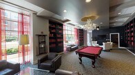 This thoughtfully designed single-floor space offers a variety of distinct areas, each with its own character and purpose. The entry area welcomes visitors with soaring ceilings, warm wood-toned double doors, polished tile flooring, and an airy, open atmosphere. A cream-upholstered sofa, pendant chandelier, and a dining table with dark chairs create an inviting, multipurpose reception zone accented by soft artwork and fresh floral arrangements.

Adjacent lounge areas feature plush neutral seating, delicate multi-light chandeliers, and black-framed French doors that flood the rooms with natural light while offering views of the outdoors. The sophisticated color palette of cream, teal, and gold carries through these spaces, with patterned carpet adding texture underfoot.

A standout feature is the stacked-stone fireplace with a rustic wood mantel, flanked by French doors opening to an exterior terrace. Nearby seating with wingback chairs creates a cozy, intimate corner perfect for conversation.

The game room delivers a striking contrast, showcasing a classic red billiards table beneath an ornate crystal chandelier with a gold canopy. Dark built-in bookshelves lined with coordinating red books, a wall-mounted cue rack, and red sheer curtains complete the dramatic, club-like aesthetic, while a lounge area with a flat-screen TV rounds out the social amenities.
