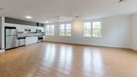 Located at 34 Main St in North Easton, Massachusetts, this two-floor space offers a thoughtful blend of contemporary design and rustic character. The main level is defined by a generously proportioned open floor plan featuring light hardwood flooring, crisp white walls, and striking exposed timber columns that lend warmth and architectural interest throughout. Track lighting runs across the ceiling, providing flexible illumination, while multiple windows along the perimeter invite abundant natural light and views of the surrounding greenery.

The kitchen on the main level is well-appointed with white cabinetry, gray countertops, a tile backsplash, and a full suite of stainless steel appliances including a refrigerator, range, dishwasher, and over-the-range microwave. A staircase connects the main floor to the upper level, where vaulted ceilings follow the roofline, creating an airy, intimate atmosphere. Upper-level rooms feature neutral carpeting and closet space, complemented by natural light from well-placed windows.

Bathrooms throughout the space are finished in a clean, modern style, featuring tile flooring, white cabinetry with warm countertops, undermount sinks, large mirrors, and a full tub-and-shower combination. The overall aesthetic balances industrial and residential sensibilities, resulting in a versatile and inviting environment.